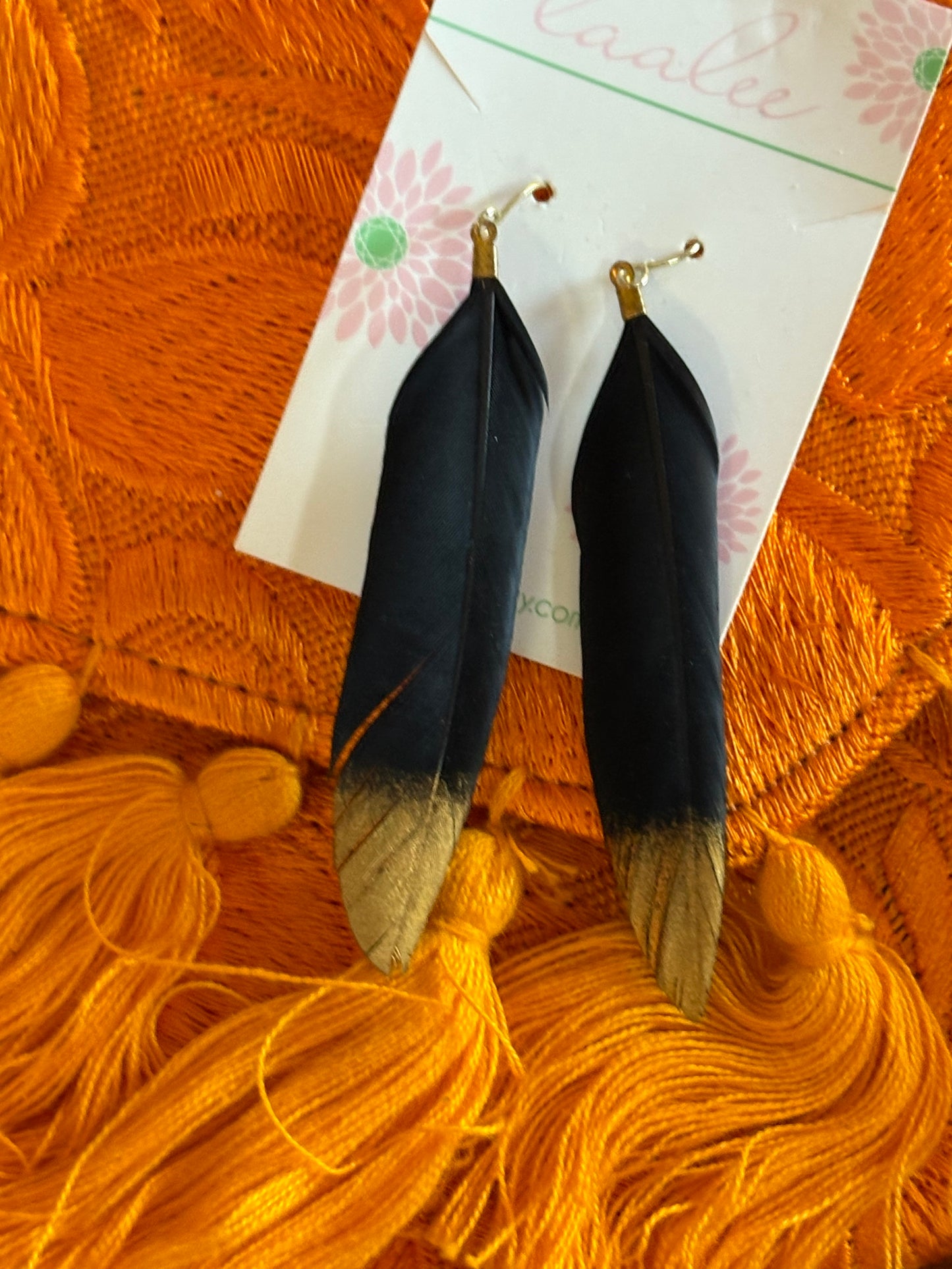Two black feathers with gold tips on a white card, placed on an orange textured surface with yellow yarn.