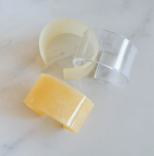 Three translucent acrylic bangle bracelets in clear, custard marble, and ivory tones displayed on a marble surface. Each bracelet is 1⅜" wide, showcasing subtle color variation and retro-inspired design.